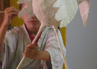 Kind mit Malerkittel bemalt einen großen, noch hauptsächlich weißen, langbeinigen Vogel, der aus Pappmaché gebastelt ist und vor dem Kind auf dem Tisch steht.